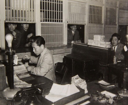은행 창구에서 은행원 두 사람이 지출 및 수납 업무를 보고 있다. 조선은행은 한국의 주요 은행이다.(48.3.26) 